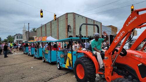 A colorful parade train pulled by a tractor, with people waving and enjoying the event on a cloudy day.