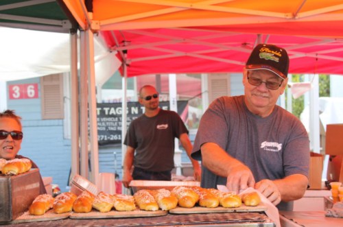 A vendor prepares hot dogs at a market stall, with colorful tents and customers in the background.