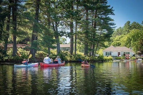Three kayakers paddle on a calm lake surrounded by trees, with a house visible in the background.