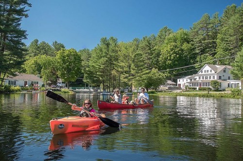 Two kayaks on a calm lake surrounded by trees, with a house in the background and people enjoying the sunny day.
