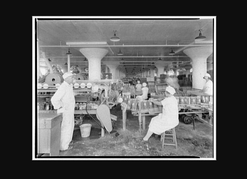 Black and white image of workers in a factory, surrounded by machinery and materials, wearing white uniforms.