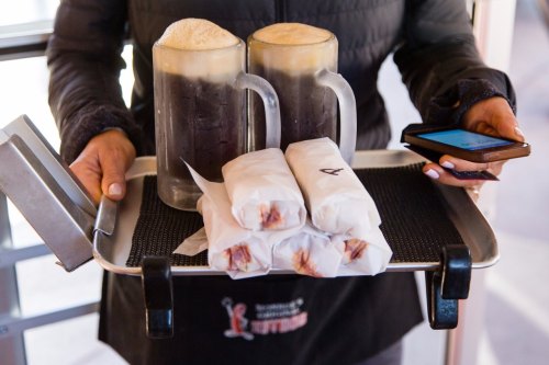 A person holds a tray with two mugs of frothy drinks and four wrapped food items, while checking a phone.
