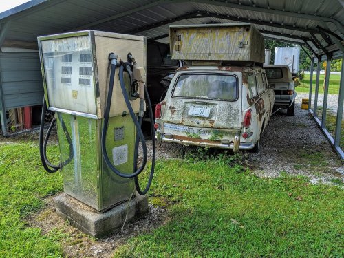 An old gas pump stands next to two vintage cars, one with a camper shell, under a metal shelter. Grass surrounds the area.