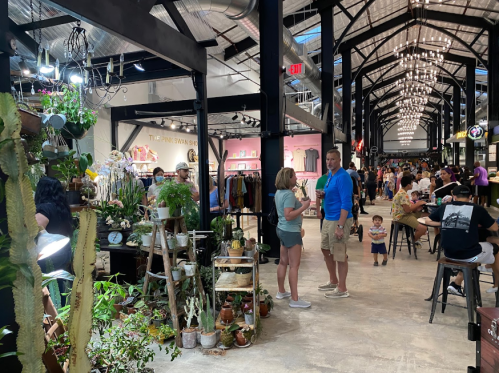 A bustling market hall with shoppers, plants for sale, and vibrant stalls under a high ceiling with lights.