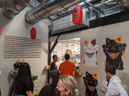 A busy shop entrance with colorful cat murals, a hanging lantern, and customers waiting in line.