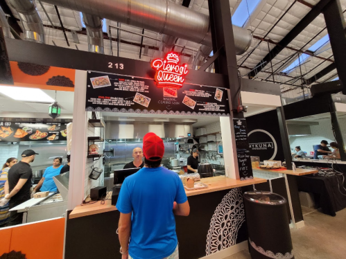 A customer stands at the counter of a food stall named "Pivogi Queen," with a menu displayed above.