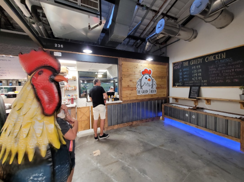 A person orders at a chicken restaurant with a large rooster statue in the foreground and a menu on the wall.