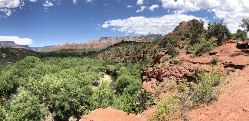 A panoramic view of a lush green valley surrounded by red rock formations and mountains under a blue sky with clouds.