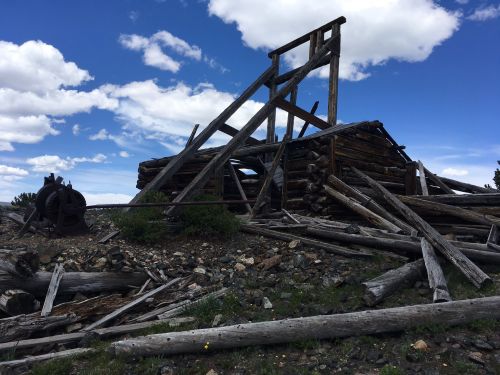 A dilapidated wooden structure with fallen beams and debris, set against a blue sky with scattered clouds.