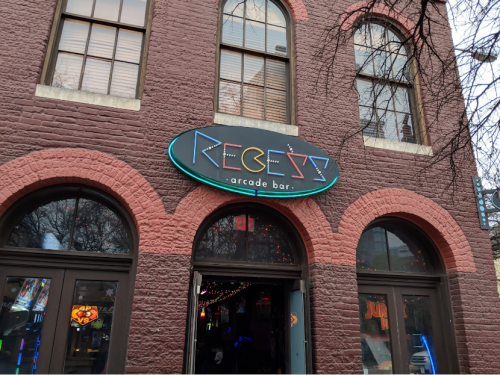 Sign for "Recess Arcade Bar" on a brick building, with neon lights and a welcoming entrance.