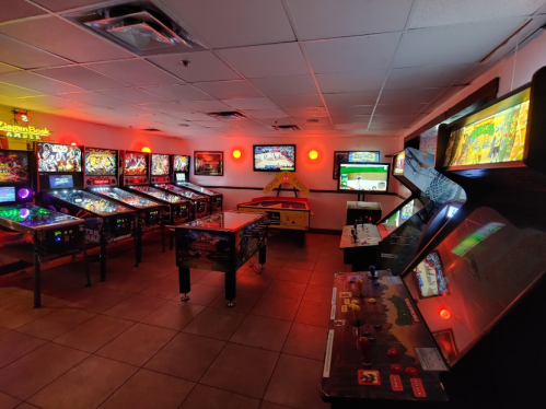 A vibrant arcade room filled with pinball machines and retro gaming consoles, illuminated by red lights.