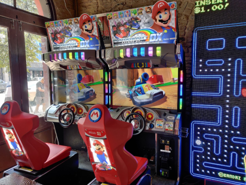 Two Mario Kart arcade machines with colorful screens and red seats, alongside a Pac-Man game in a lively arcade setting.