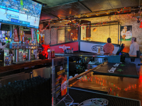 A lively bar scene with a basketball game on TV, colorful lights, and patrons enjoying drinks at the counter.