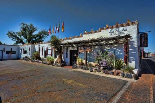 A charming white building with flags, plants, and a sign reading "La Posta de Mesilla" in a sunny outdoor setting.