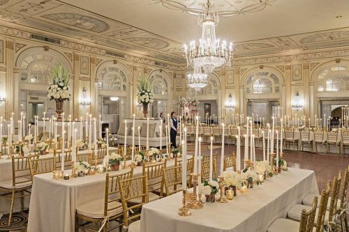 Elegant banquet hall with long tables, golden chairs, candles, and floral arrangements under a chandelier.