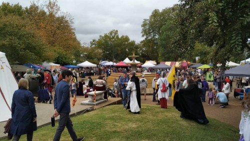 A lively outdoor festival scene with people in medieval attire, tents, and a fountain surrounded by trees.