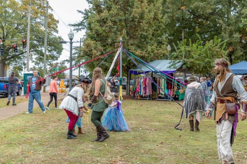 A lively outdoor scene with people dancing around a maypole, colorful ribbons, and vendors in the background.