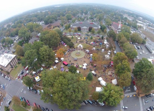 Aerial view of a bustling outdoor festival with tents, people, and trees in a park-like setting.