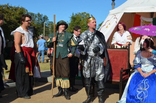 A group of people in medieval costumes, including a knight, a jester, and a woman in a blue gown, at an outdoor event.