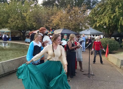 A group of people in colorful costumes perform at an outdoor event, with tents and trees in the background.