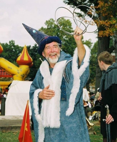 A smiling wizard in a blue robe and hat performs with metal rings at a festive outdoor event.
