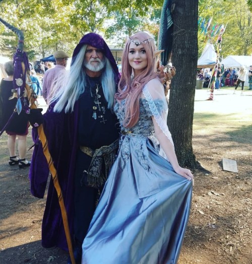 A man in a wizard costume and a woman in an elegant gown pose together at an outdoor festival.