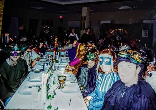 A festive gathering with people wearing colorful masks, seated at a long table decorated for a celebration.