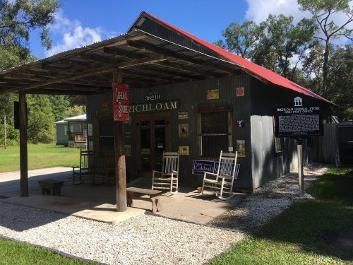 A rustic general store with a red roof, rocking chairs, and a historical marker in a green, wooded area.