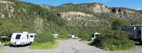 A scenic view of a campground with RVs surrounded by greenery and mountains under a clear blue sky.