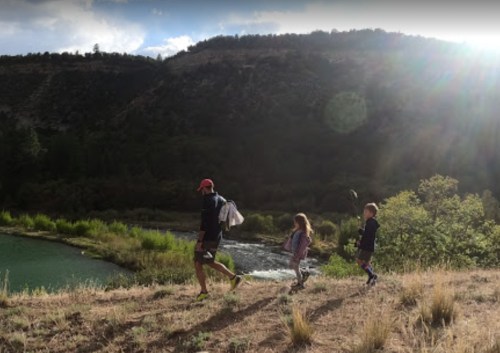 A person and two children walk along a riverbank, surrounded by greenery and mountains under a partly cloudy sky.