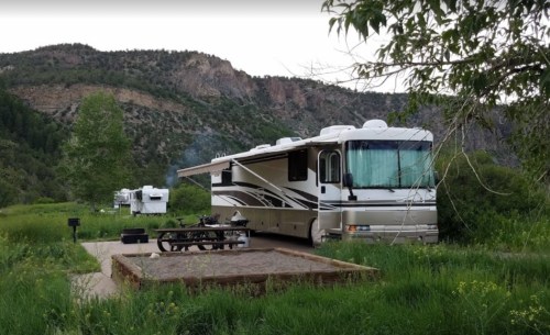 A parked RV in a green campsite surrounded by trees and mountains, with a fire pit in the foreground.