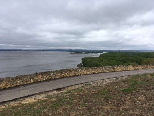 A winding road along a riverbank, with lush greenery and overcast skies in the background.