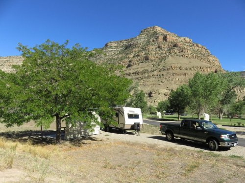 A camper and truck parked near a tree, with a rocky hill in the background under a clear blue sky.