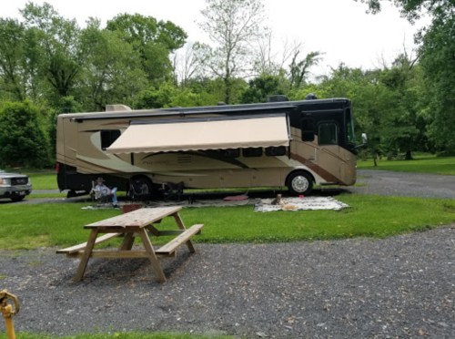 A large RV parked on a grassy area with a picnic table nearby and trees in the background. An awning is extended.