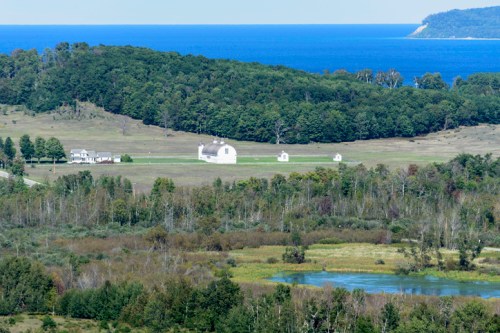A scenic view of rolling hills, a calm blue lake, and distant ocean, with white buildings dotting the landscape.