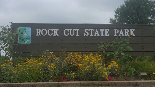 Sign for Rock Cut State Park surrounded by colorful flowers and greenery under a cloudy sky.