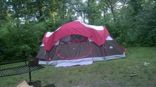 A red and brown tent set up in a grassy area surrounded by trees and greenery.
