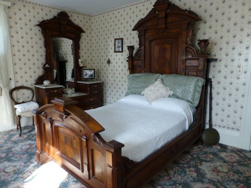 A vintage bedroom featuring a wooden bed, floral wallpaper, and antique furniture, creating a cozy atmosphere.