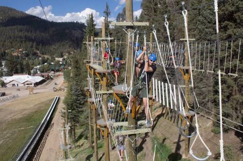 A group of people navigate a high ropes course among trees, wearing helmets and harnesses, with a scenic backdrop.