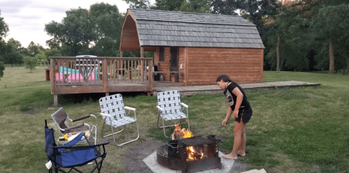 A girl stands by a campfire in front of a wooden cabin, with chairs nearby and a grassy area in the background.