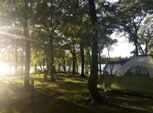 A serene campsite with a green tent surrounded by trees, sunlight filtering through, and a view of a lake.