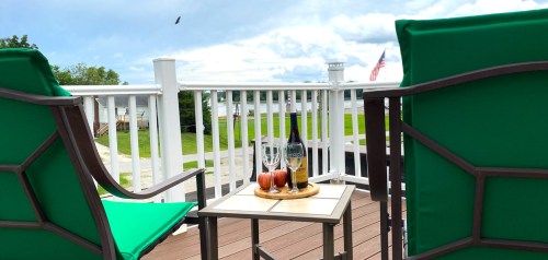 Two green chairs on a deck with a table holding a bottle and glasses, overlooking a scenic view with an American flag.