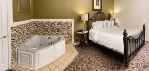 A cozy hotel room featuring a bed, decorative lamps, and a corner jacuzzi tub with tiled walls.
