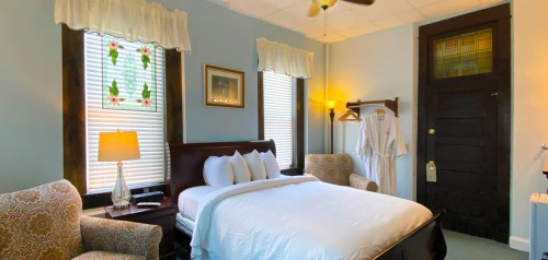 Cozy bedroom with a queen bed, armchairs, lamp, and a robe hanging on a rack, featuring light blue walls and natural light.