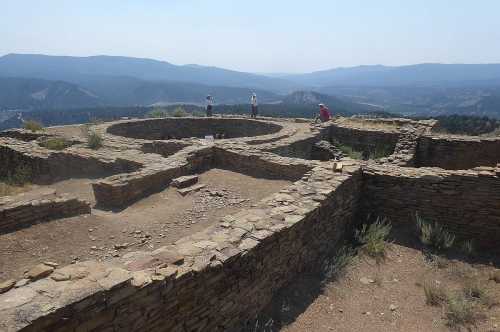 Ancient stone ruins on a hillside with visitors exploring, surrounded by mountains and a clear blue sky.