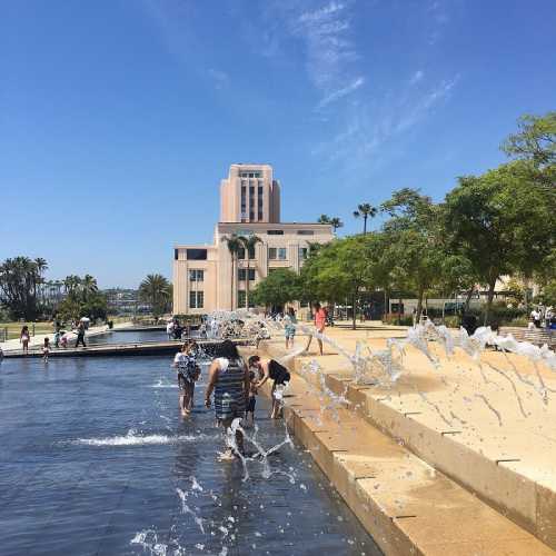 People play in a fountain at a sunny park, with palm trees and a building in the background.