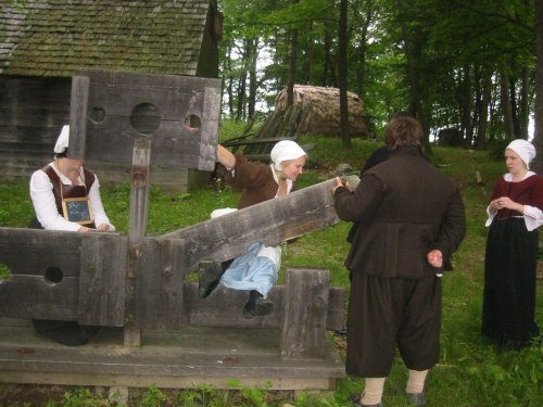 Historical reenactors demonstrate a stocks punishment in a wooded area, with traditional clothing and a rustic backdrop.