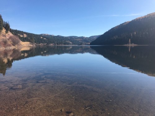 A serene lake reflecting mountains and trees under a clear blue sky. The water is calm and transparent.