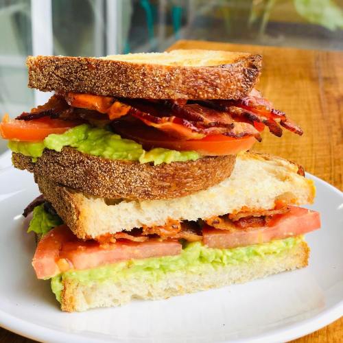 A stacked sandwich with crispy bacon, fresh tomato, and avocado spread between toasted bread slices on a white plate.