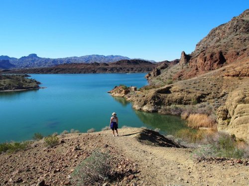 A person walks along a trail by a serene lake, surrounded by rocky hills and clear blue skies.